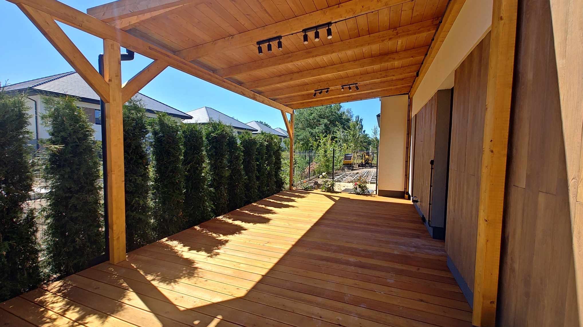 Zadaszony taras z desek, oświetlony punktowymi lampami, z widokiem na ogród i domy sąsiadów, otoczony tujami.