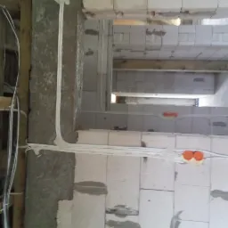 Kable elektryczne prowadzone wzdłuż surowych ścian z bloczków betonowych, widoczne pomarańczowe puszki instalacyjne i drewniane elementy konstrukcyjne.