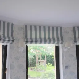 Trzy okna z szaro-białymi roletami rzymskimi w paski, ściana w kwiatowy wzór, widok na zielony ogród z domkiem dla ptaków.