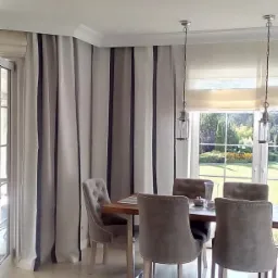 Jadalnia z zasłonami w pionowe pasy w odcieniach szarości, roletą rzymską w oknie i szarymi krzesłami tapicerowanymi wokół drewnianego stołu.