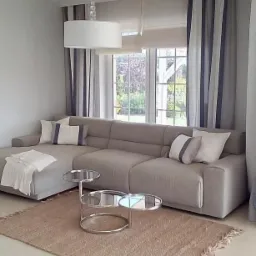 Jasny salon z narożnikiem, dywanem z frędzlami i stolikiem kawowym z trzema blatami, okno z roletą rzymską i zasłonami w pasy.