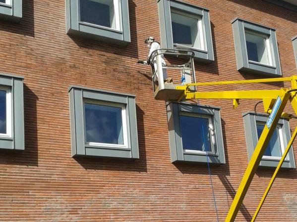 Pracownik w kombinezonie ochronnym na podnośniku koszowym czyści elewację budynku z cegły, widoczne ślady czyszczenia na murze.