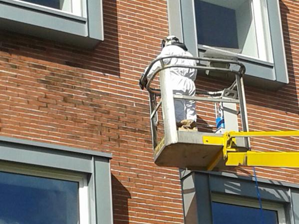 Pracownik w kombinezonie ochronnym na podnośniku koszowym czyści ceglaną elewację budynku, widoczne ślady zabrudzeń przed czyszczeniem.