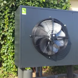 Szara jednostka zewnętrzna z wentylatorem na tle zielonej ściany z bluszczu i metalowej bramy.