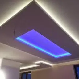 Sufit podwieszany z oświetleniem LED: prostokątna wnęka z niebieskim światłem w centralnej części, obramowana białym światłem LED, widoczne gładkie wykończenie i fragment ściany z oknem.