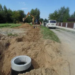 Betonowy krąg studzienny w wykopie, w tle pracujący koparka, biały bus i dwuosobowa ekipa przyłącza na terenie budowy w słoneczny dzień.
