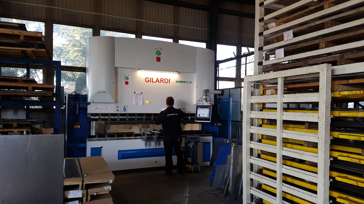 Pracownik obsługuje białą prasę krawędziową Gilardi Olympia 3.12 w hali produkcyjnej, widoczne metalowe półki z arkuszami blachy i paletami.