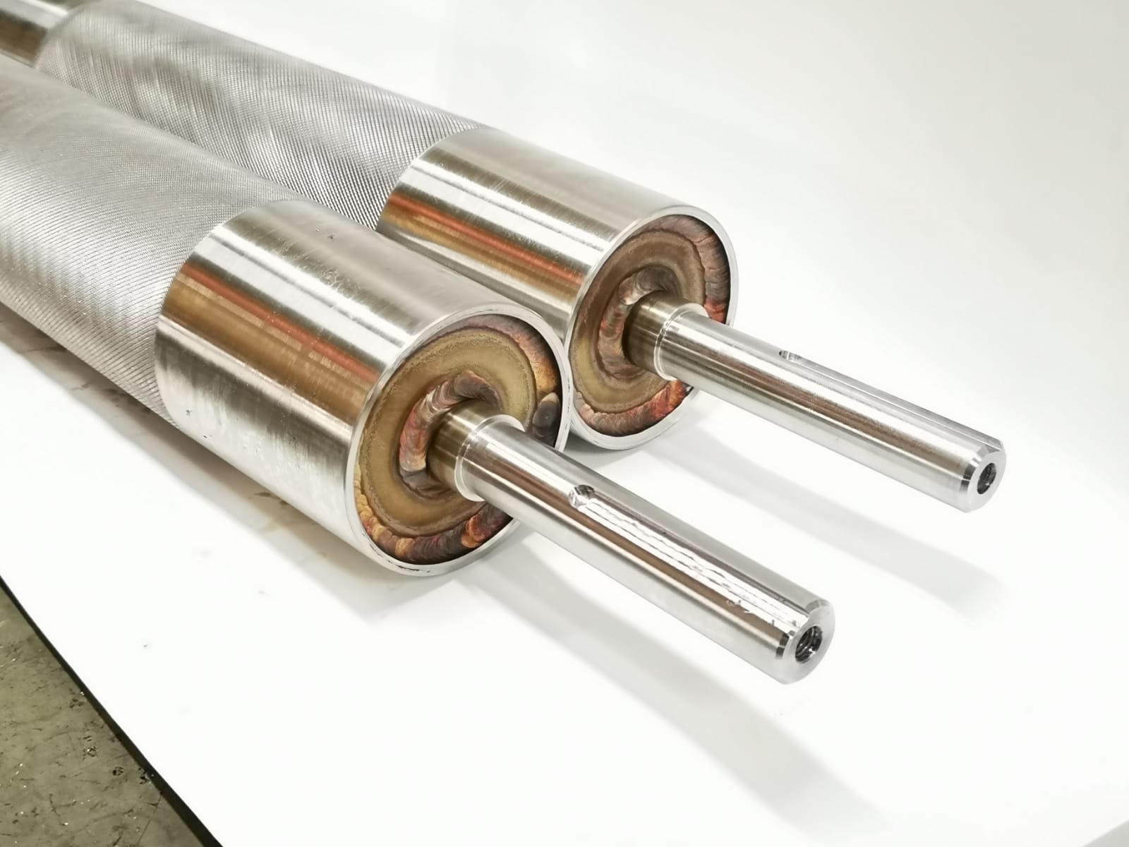 Dwa metalowe wałki z teksturowaną powierzchnią i przyspawanymi końcówkami, widoczne ślady spawania, na białym tle.