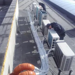 Instalacja rzędu klimatyzatorów na płaskim dachu budynku, widoczne pomarańczowe rury kanalizacyjne i wiązka białych kabli, dwóch pracowników w trakcie montażu.