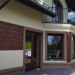 P.U. ELEWATOR MARIUSZ KOWALCZYK - Dwukondygnacyjny dom z beżową elewacją, drewnopodobnym panelem na ścianie i balkonem z ozdobną balustradą, widok z zewnątrz w słoneczny dzień.