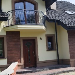 P.U. ELEWATOR MARIUSZ KOWALCZYK - Jasnożółty dom jednorodzinny z ciemnobrązową stolarką okienną i drzwiową, balkonem z kutą balustradą, elewacją z elementami imitującymi drewno i ciemną dachówką.