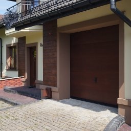 P.U. ELEWATOR MARIUSZ KOWALCZYK - Elewacja domu jednorodzinnego z brązową bramą garażową i elementami dekoracyjnymi imitującymi drewno, widoczne panele solarne na dachu sąsiedniego budynku.