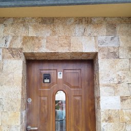 P.U. ELEWATOR MARIUSZ KOWALCZYK - Wejście do domu z elewacją z kamienia naturalnego w odcieniach beżu i brązu, brązowe drzwi z małym okienkiem i domofonem.