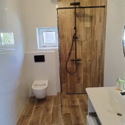 Nowoczesna łazienka z drewnopodobnymi płytkami na ścianie prysznica i podłodze, białe płytki na pozostałych ścianach, wisząca toaleta i umywalka z szafką, czarna armatura prysznicowa.