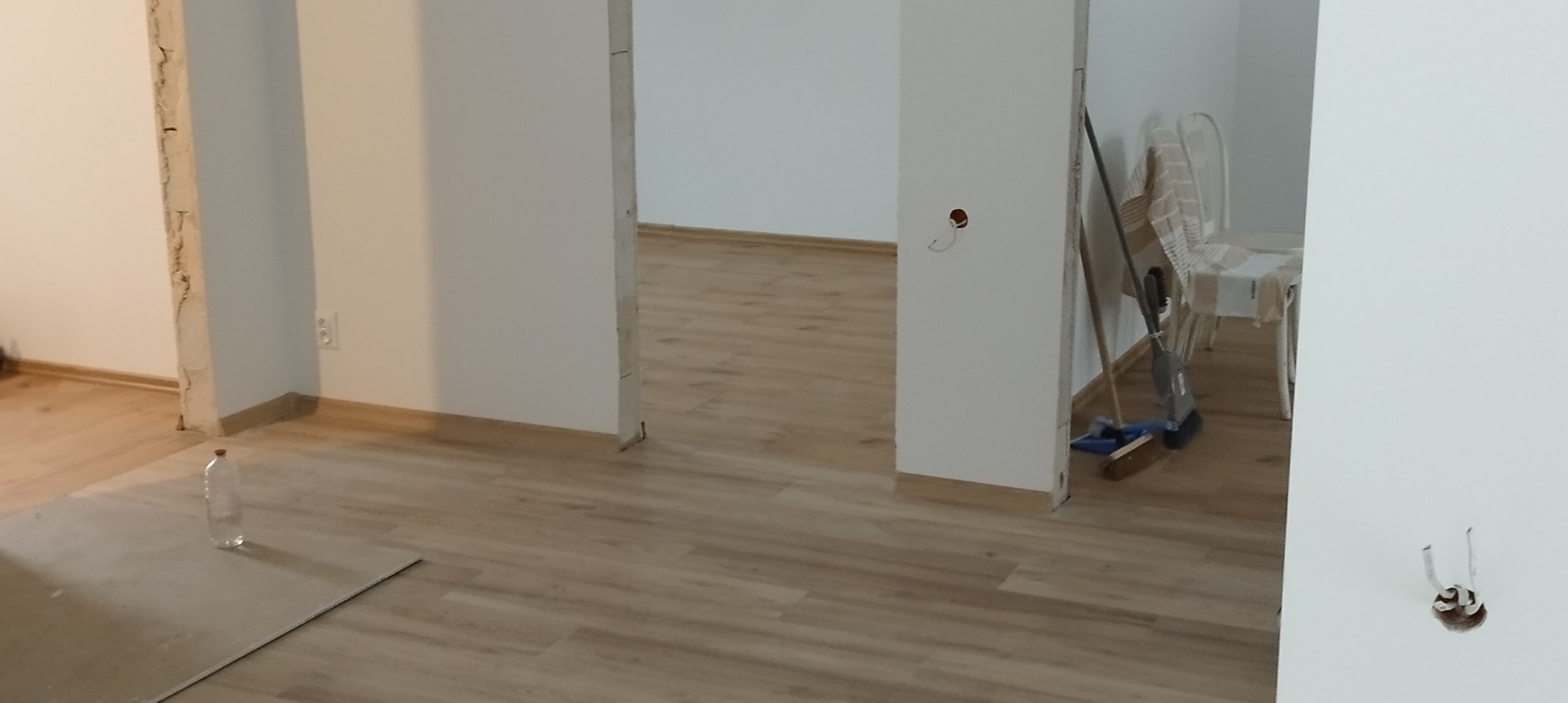 Wnętrze mieszkania w trakcie prac wykończeniowych, widoczne fragmenty ścian z instalacją elektryczną, podłoga z paneli oraz narzędzia pracy i butelka wody.