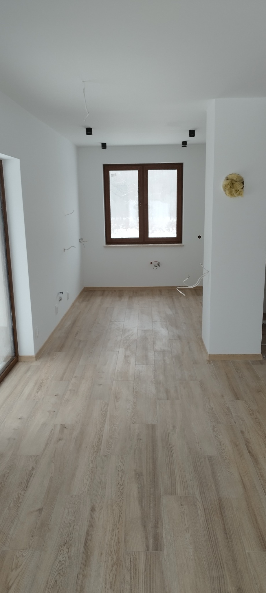 Wnętrze po remoncie z parkietem w odcieniach beżu, białymi ścianami, brązowymi oknami i wystającymi kablami elektrycznymi, przygotowane do dalszych prac wykończeniowych.