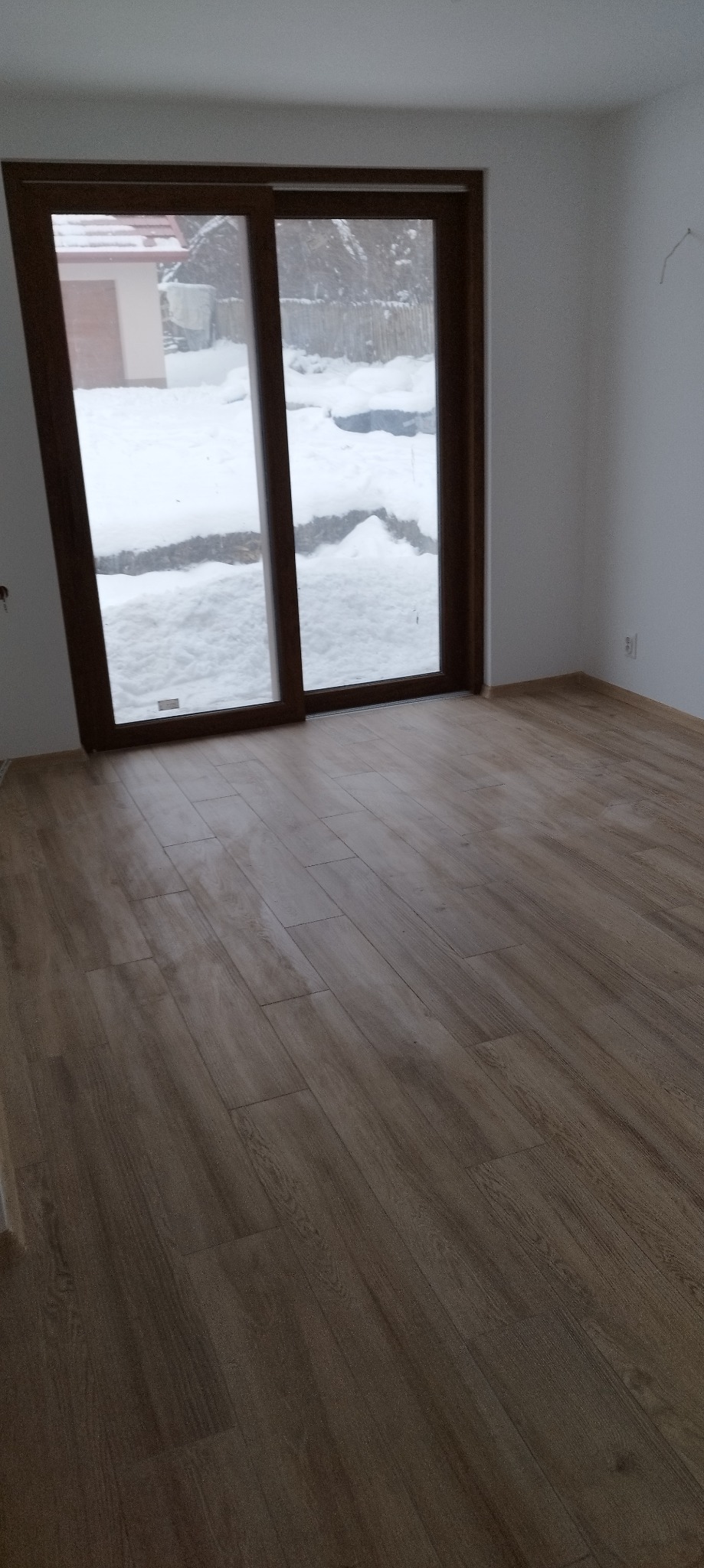 Jasnobrązowa podłoga imitująca drewno w pustym pokoju z oknem balkonowym, za którym widać śnieg.