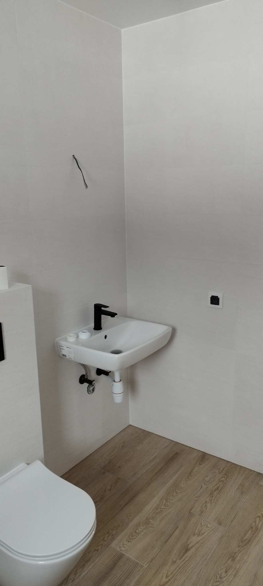 Nowoczesna, minimalistyczna łazienka z białą armaturą, drewnopodobną podłogą i czarną baterią umywalkową; widoczne niedokończone podłączenie elektryczne.