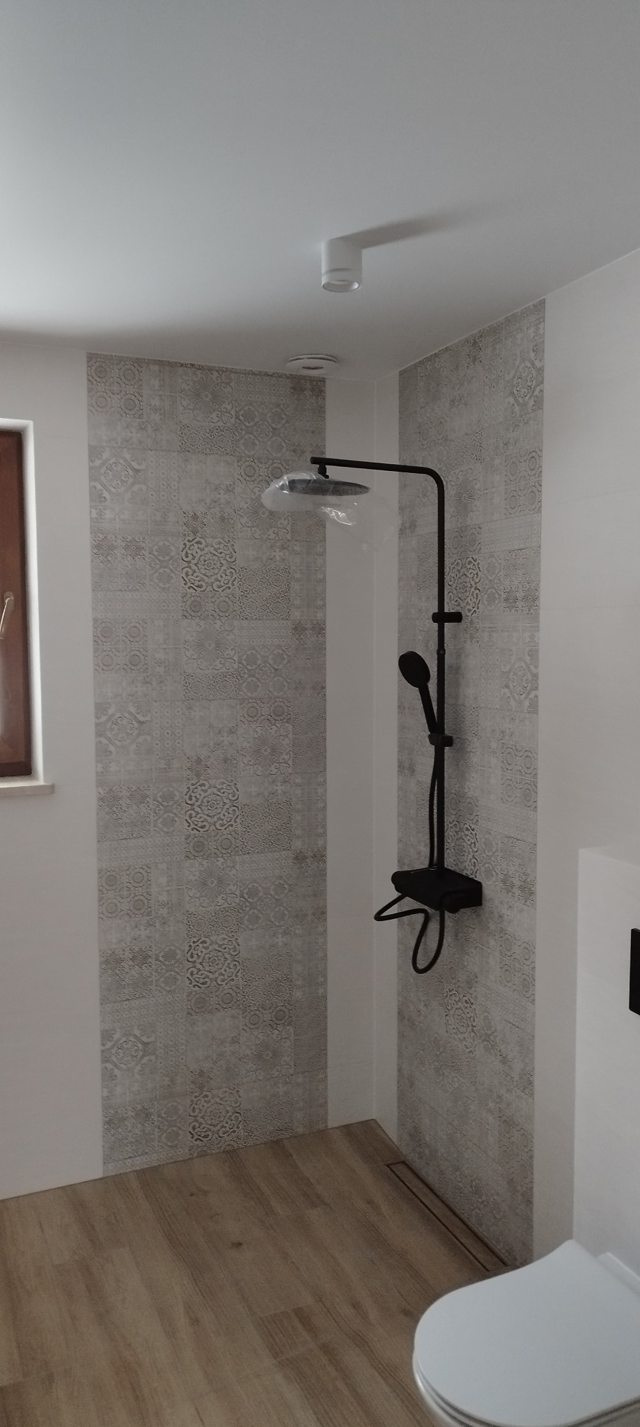 Nowoczesna kabina prysznicowa z czarną armaturą i dekoracyjnymi płytkami ściennymi w odcieniach szarości, z drewnopodobną podłogą i odpływem liniowym.