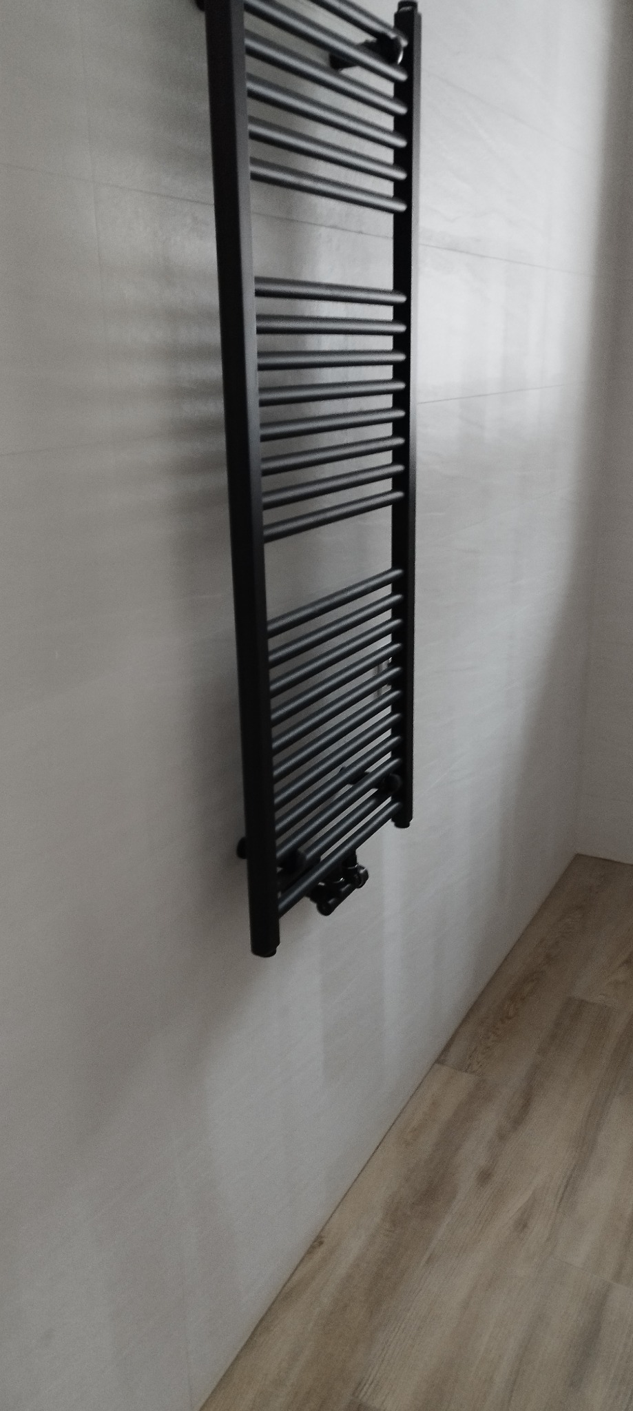 Czarny, drabinkowy grzejnik łazienkowy zamontowany na jasnej, płytkowej ścianie, z widoczną drewnopodobną podłogą w tle.