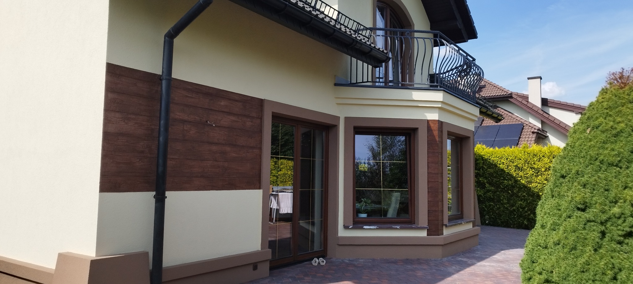 Dwukondygnacyjny dom z beżową elewacją, drewnopodobnym panelem na ścianie i balkonem z ozdobną balustradą, widok z zewnątrz w słoneczny dzień.