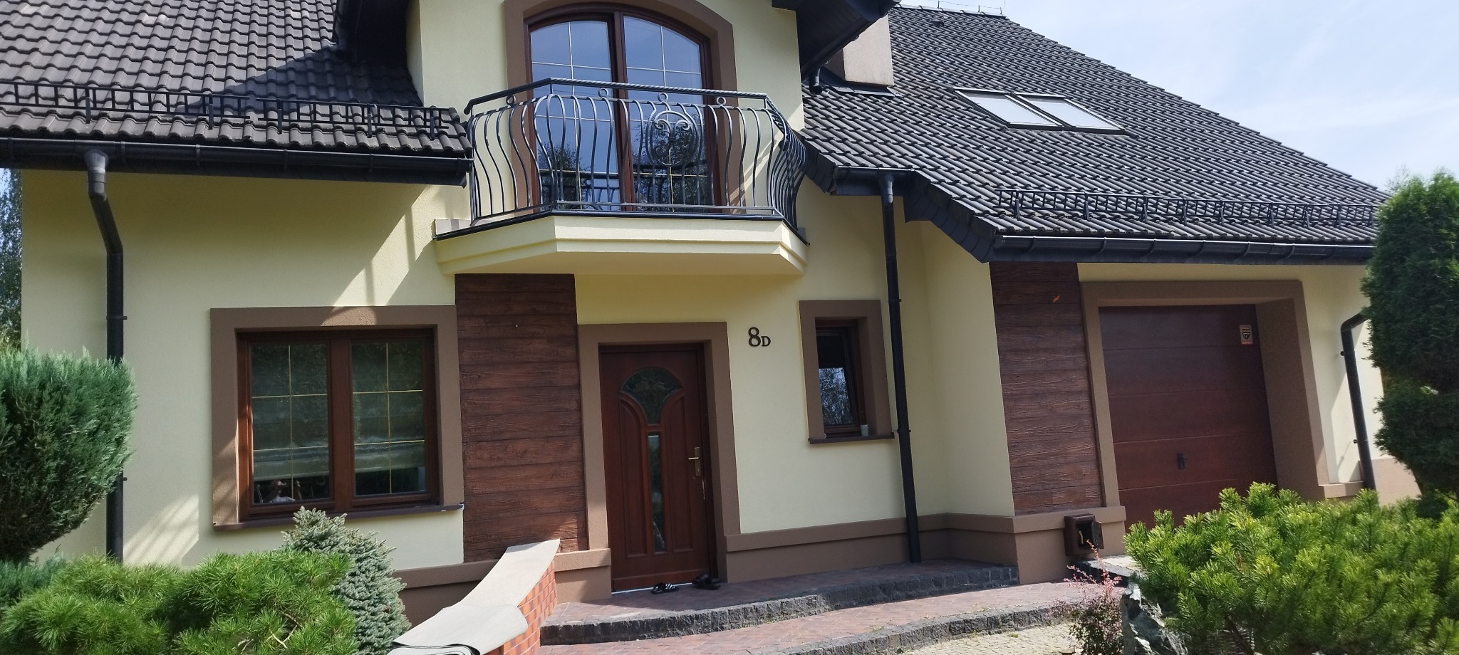 Jasnożółty dom jednorodzinny z ciemnobrązową stolarką okienną i drzwiową, balkonem z kutą balustradą, elewacją z elementami imitującymi drewno i ciemną dachówką.