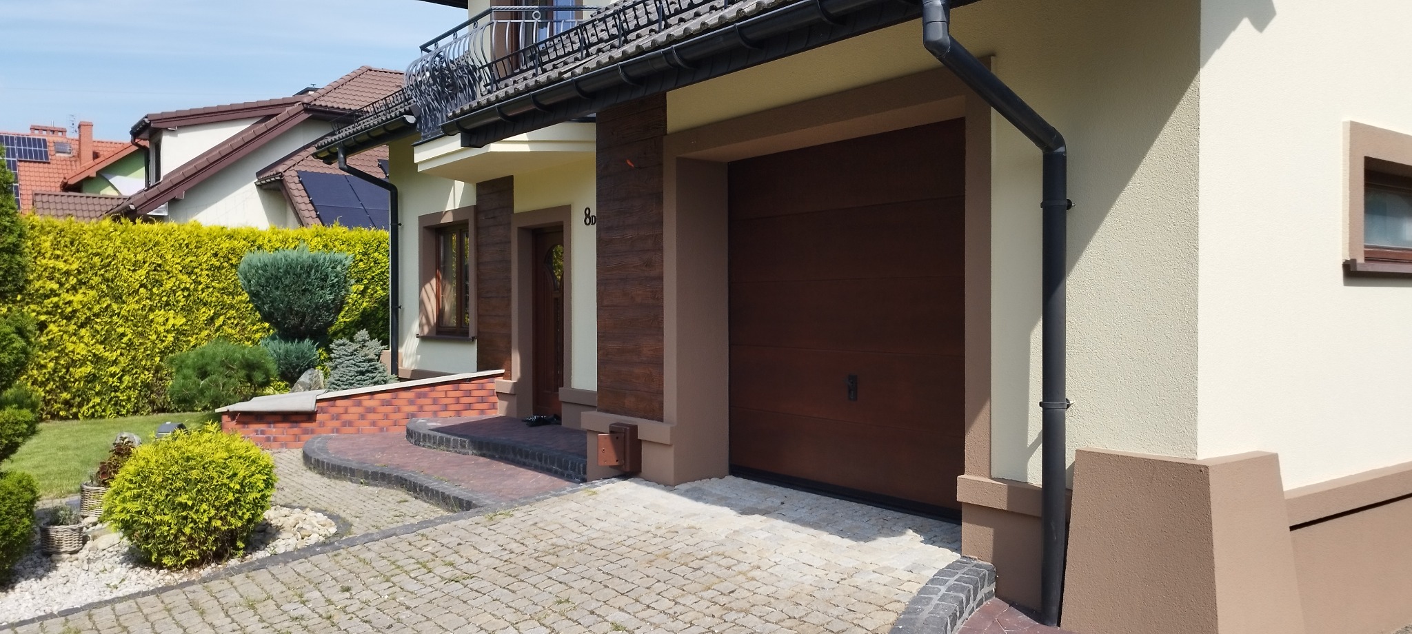 Elewacja domu jednorodzinnego z brązową bramą garażową i elementami dekoracyjnymi imitującymi drewno, widoczne panele solarne na dachu sąsiedniego budynku.