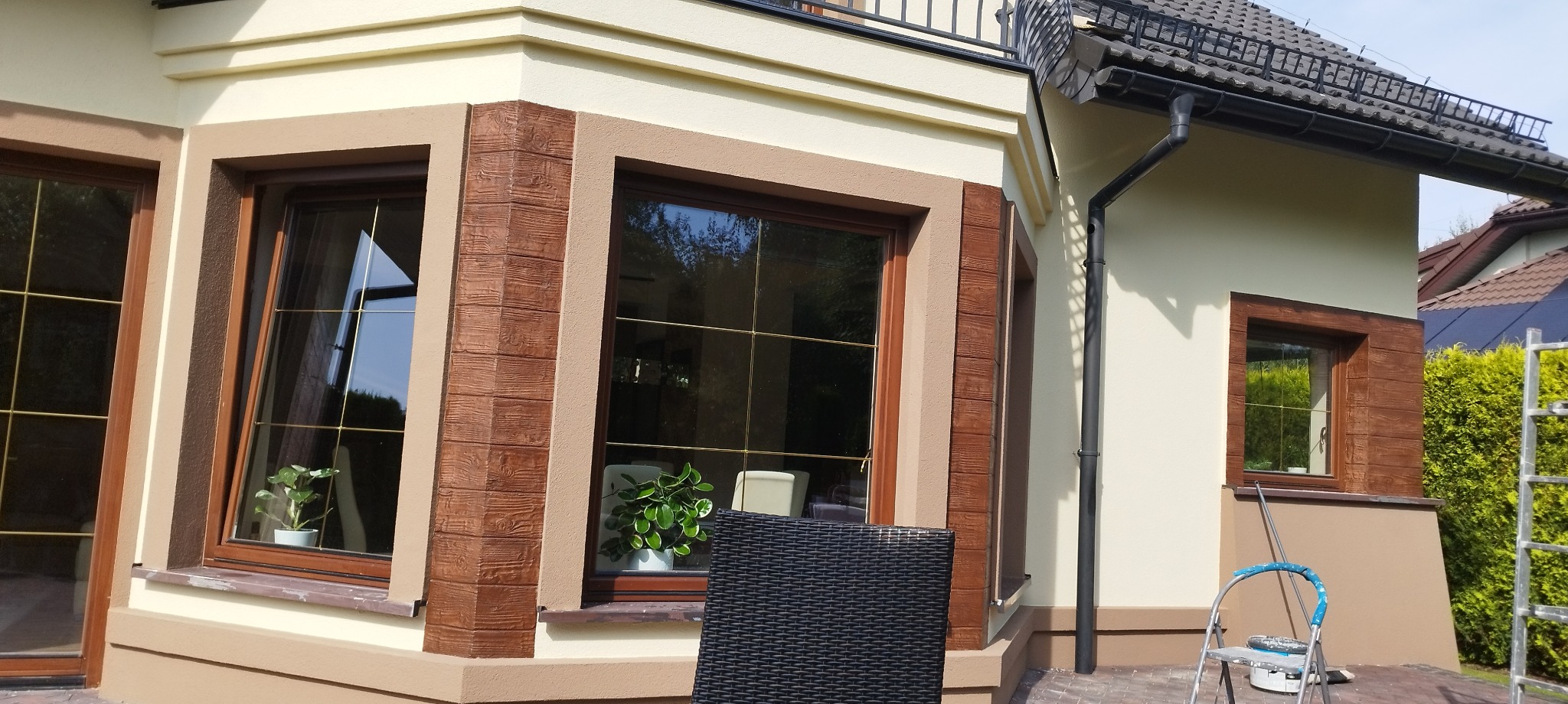 Elewacja domu z oknami o brązowych ramach, wykończona dekoracyjnym tynkiem imitującym drewno, widoczne rusztowanie i drabina.