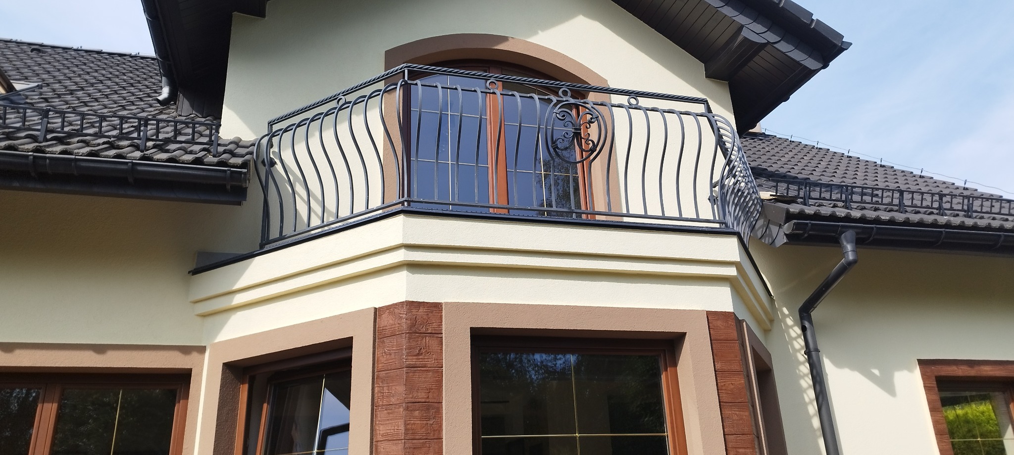 Elewacja domu z beżową ścianą, brązowymi detalami okiennymi i ozdobnym, czarnym balkonem z metalowymi, zakrzywionymi prętami.