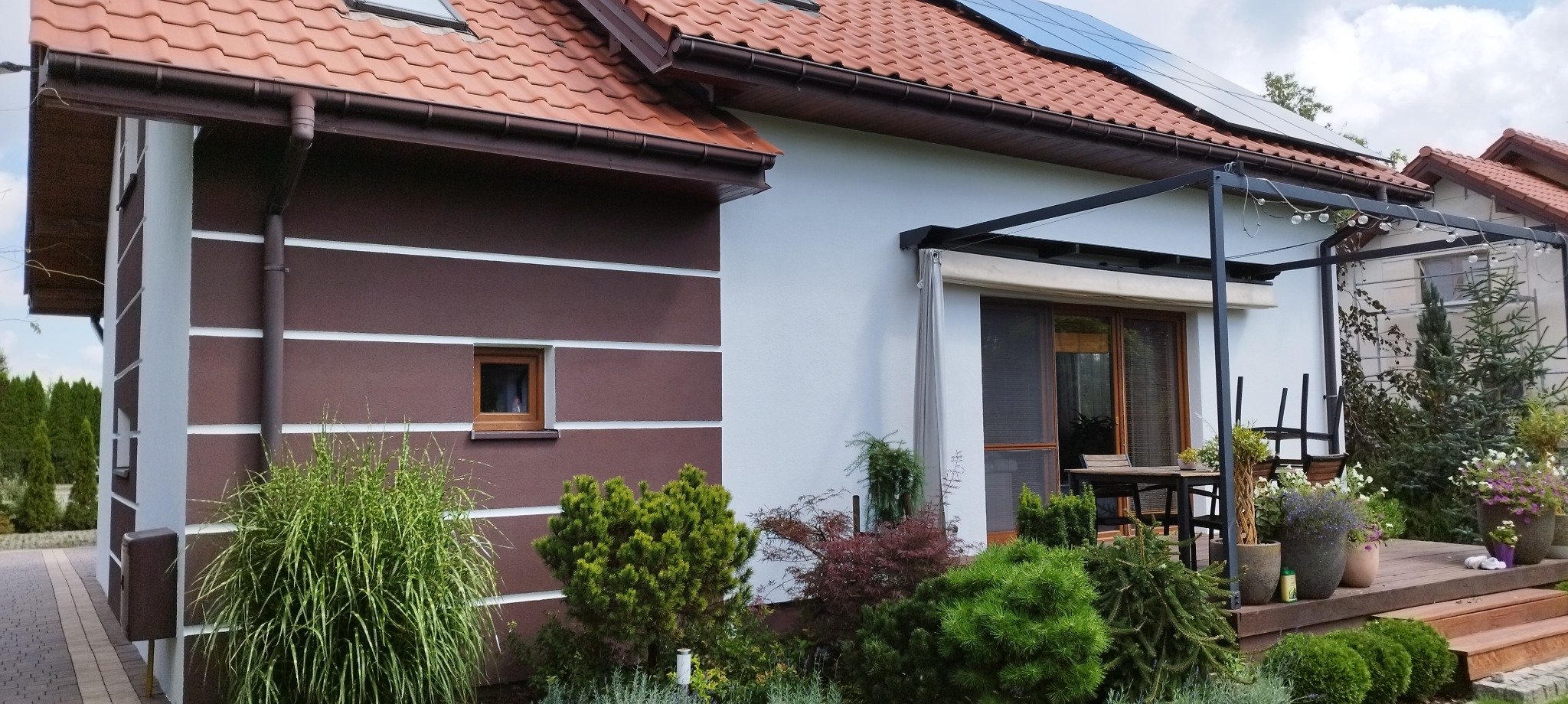 Dwukolorowa elewacja domu z brązowymi poziomymi pasami i białymi ścianami, czerwona dachówka, panele słoneczne i drewniany taras z pergolą.