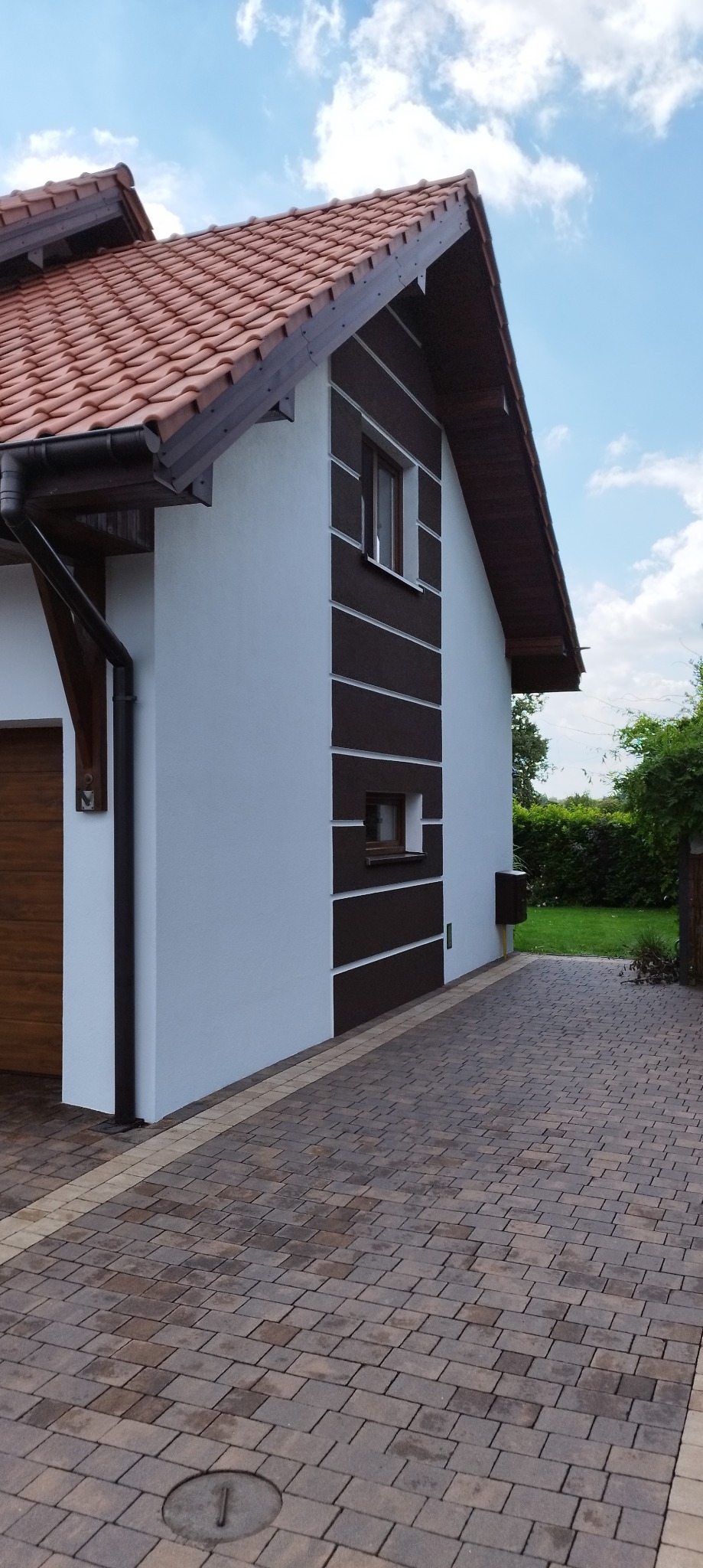 Dwukondygnacyjny dom z białą elewacją i brązowymi akcentami, widoczne okna, dachówka i brukowana ścieżka.