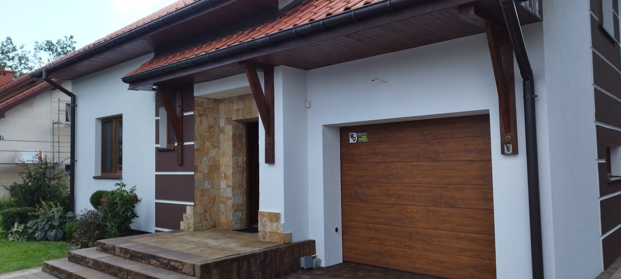 Biała elewacja domu z brązowymi akcentami, drewnianą bramą garażową i wejściem wykończonym kamieniem dekoracyjnym, widoczne schody wejściowe.