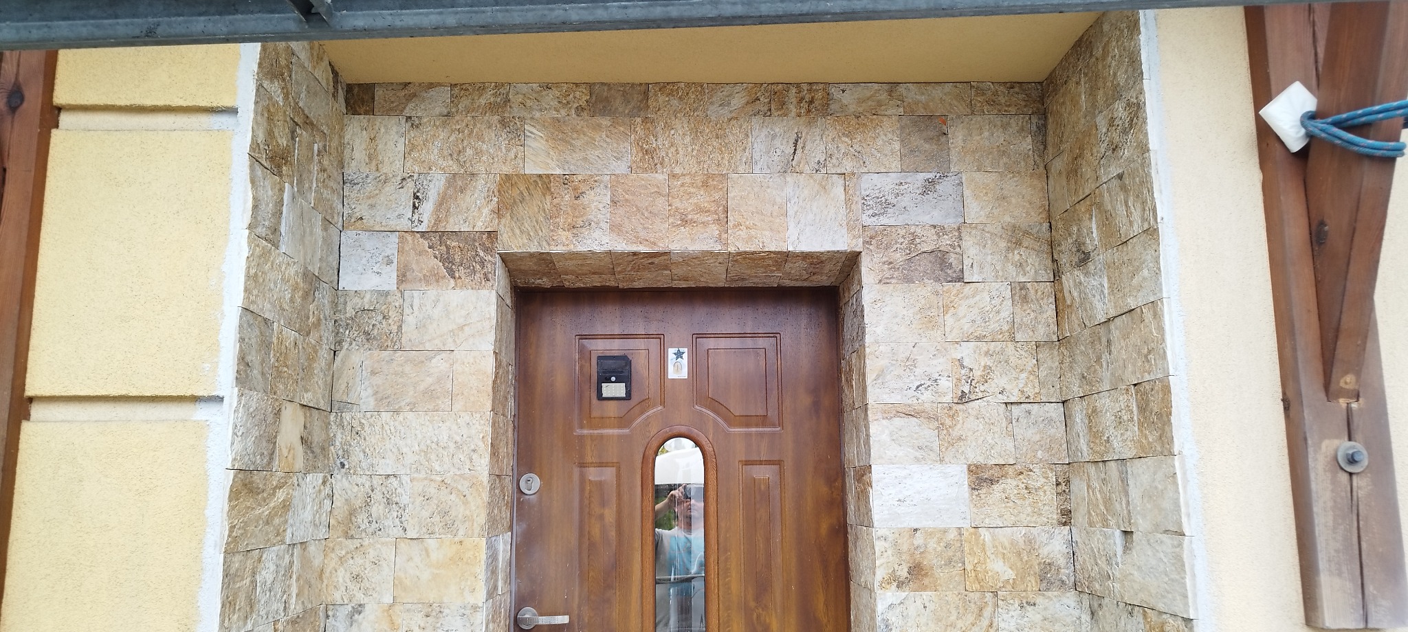 Wejście do domu z elewacją z kamienia naturalnego w odcieniach beżu i brązu, brązowe drzwi z małym okienkiem i domofonem.