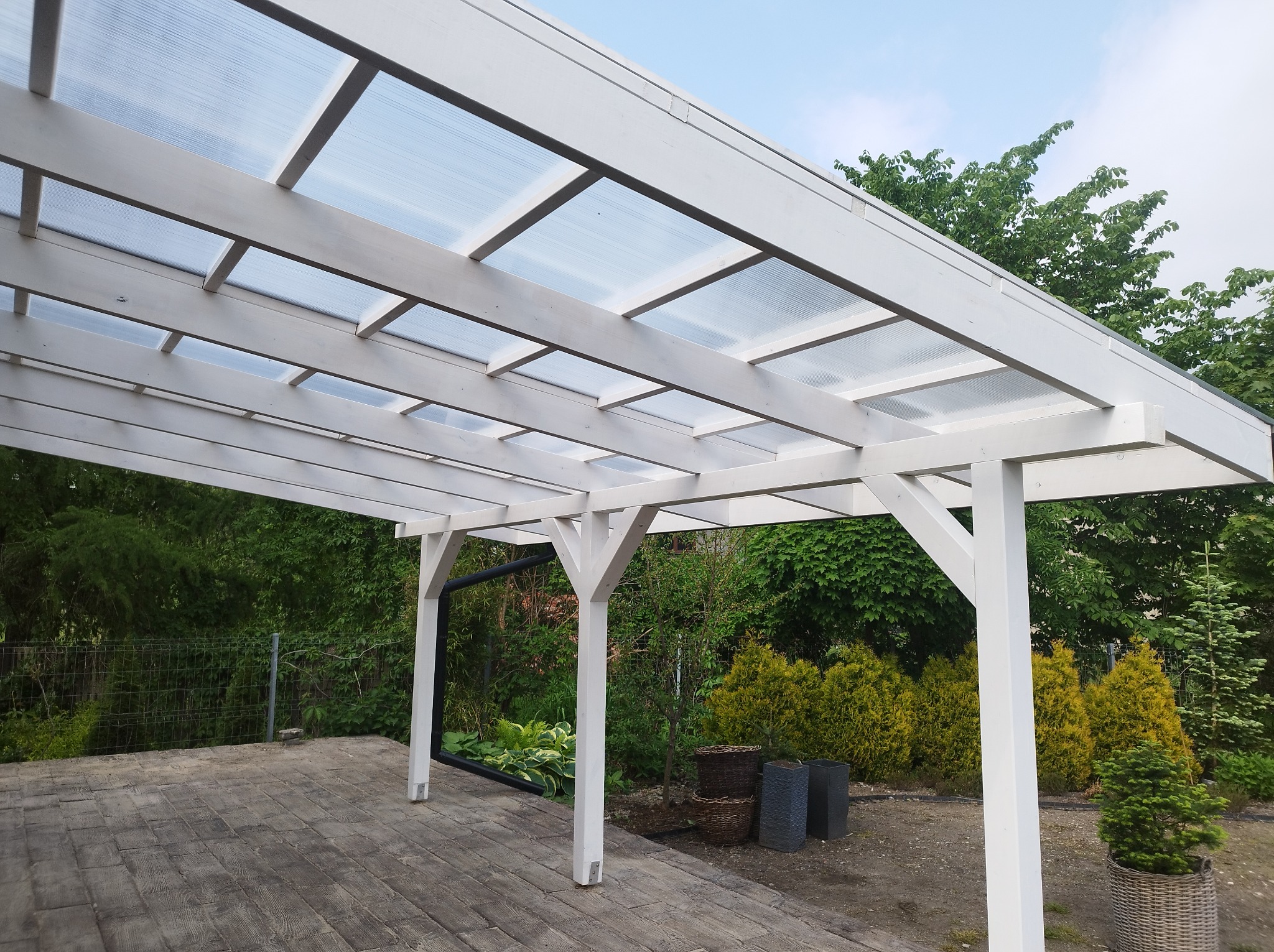 Biała drewniana pergola z przezroczystym zadaszeniem na betonowej kostce, otoczona zielenią ogrodu.