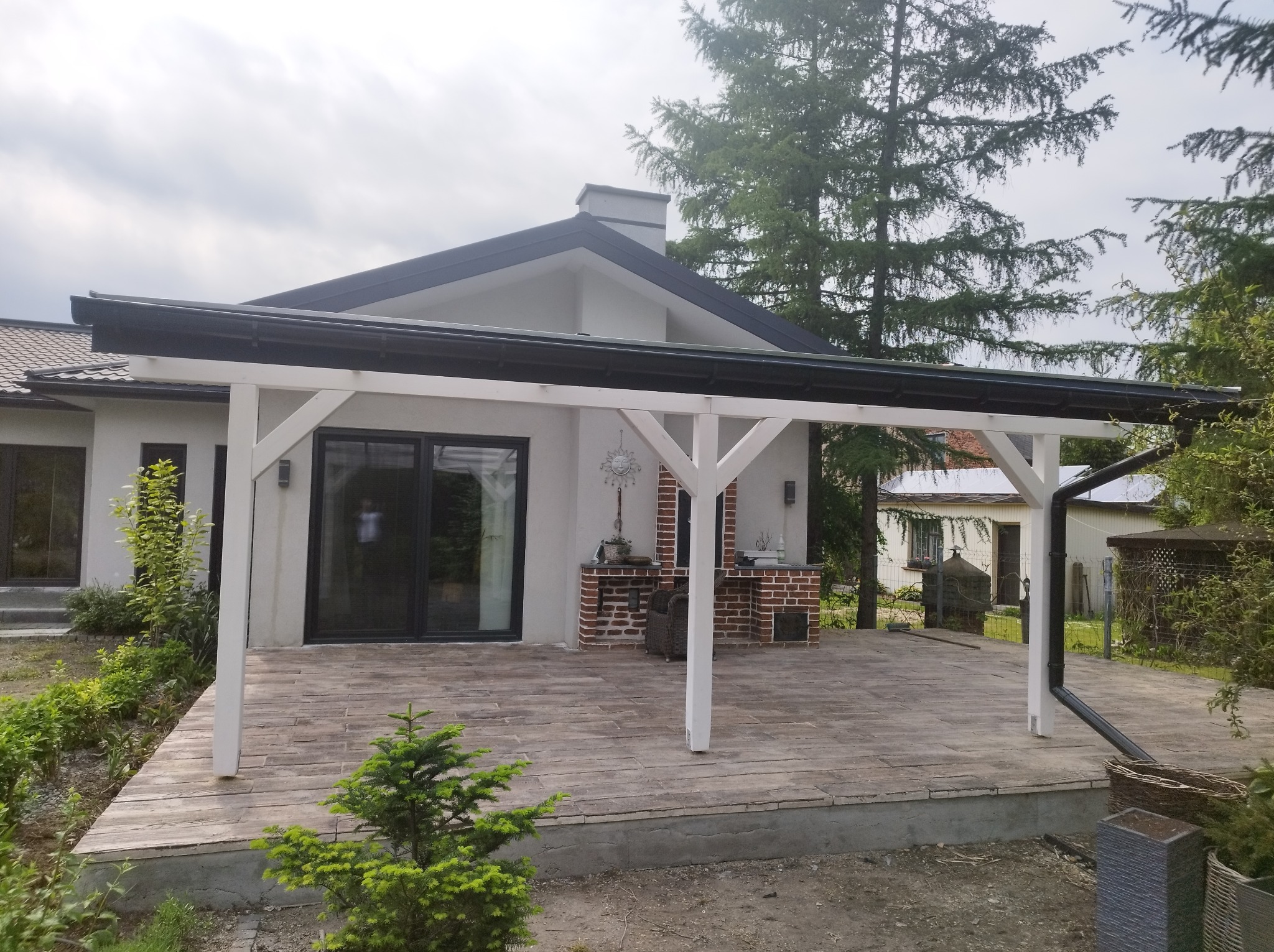 Zadaszony taras z białymi drewnianymi słupami i czarnym dachem, wyłożony płytami imitującymi drewno, z murowanym grillem w tle i zadbaną zielenią.