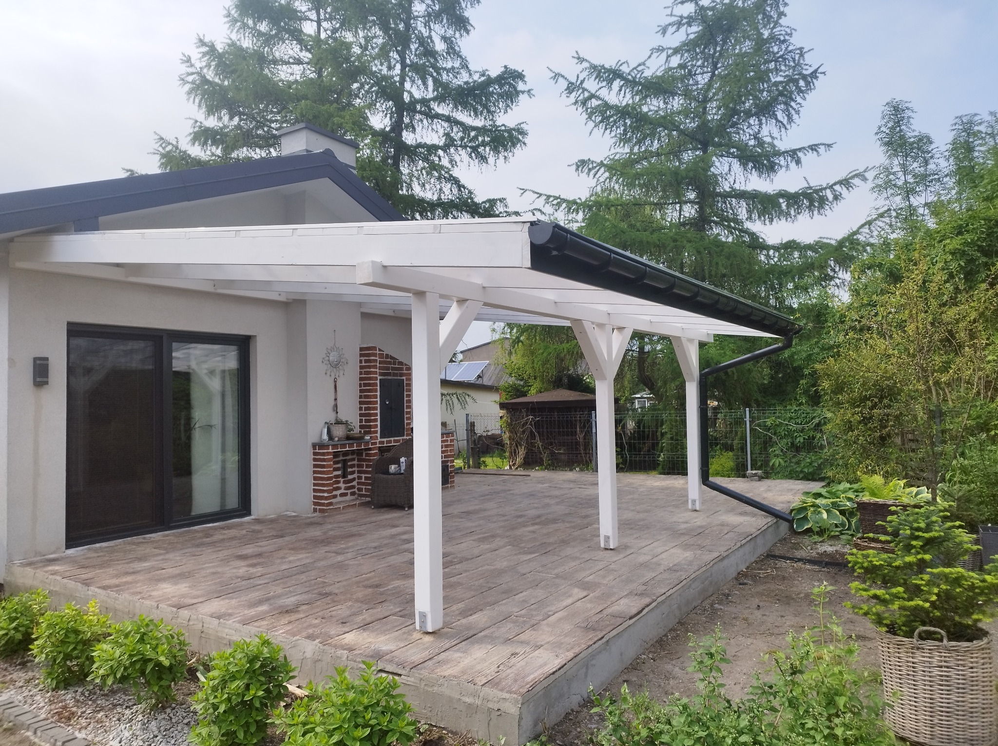 Zadaszony taras z drewnianą podłogą i białymi słupami, przylegający do nowoczesnego domu z ceglanym grillem, otoczony zielenią.