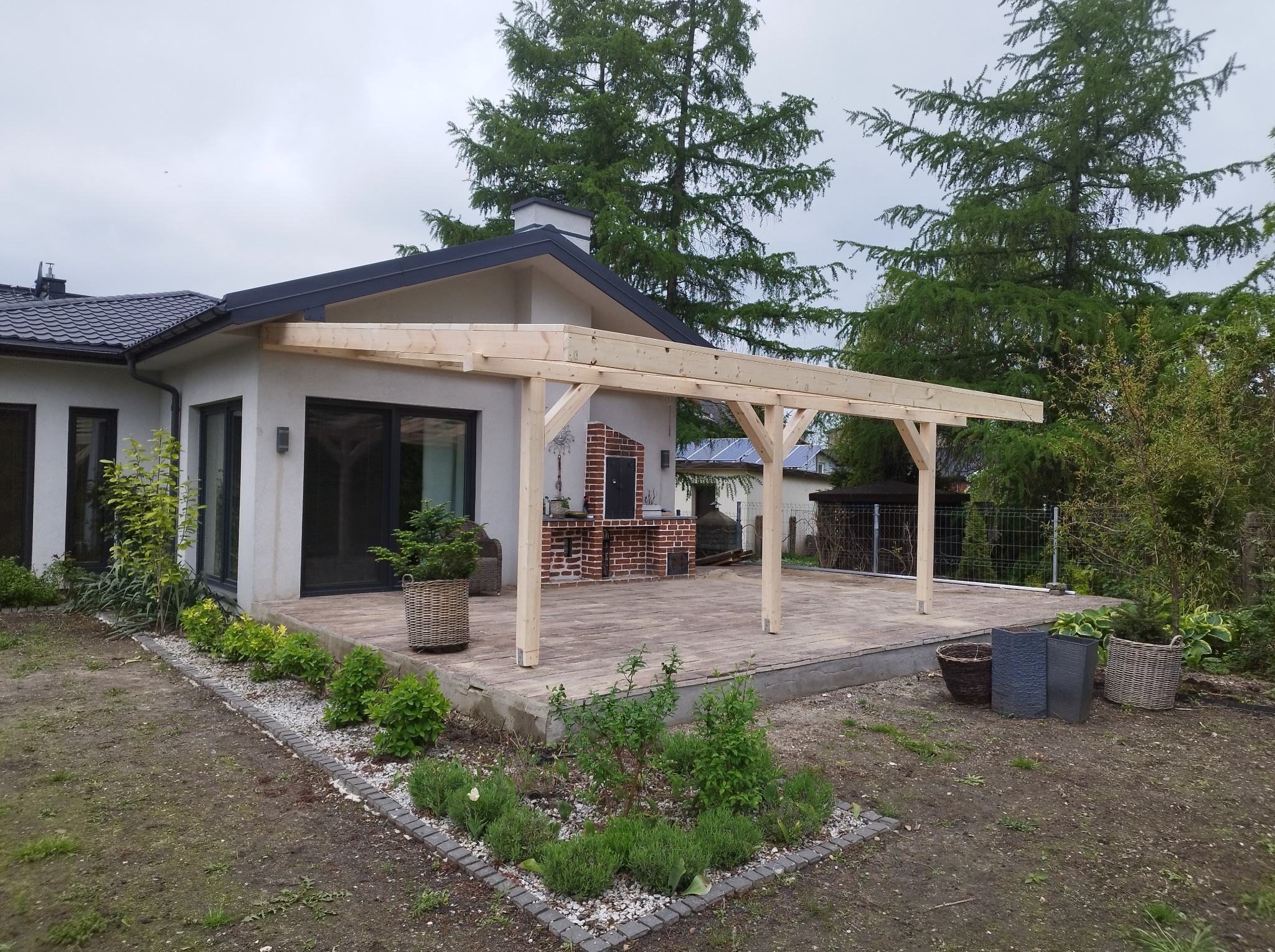 Konstrukcja drewnianej pergoli w trakcie budowy, przylegająca do domu z szarą elewacją i czarnym dachem, z widocznym murowanym grillem w tle.