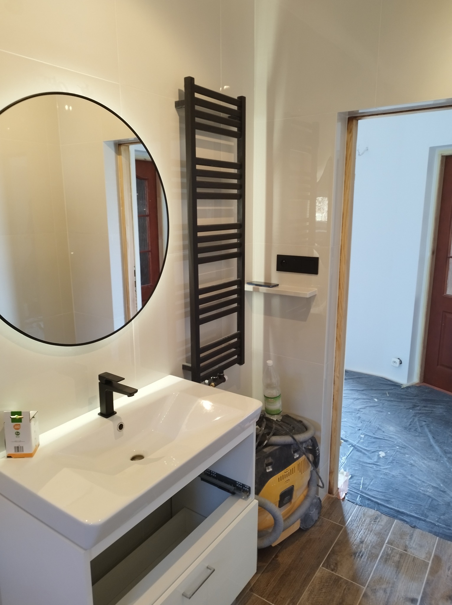 Nowoczesna łazienka z okrągłym lustrem z podświetleniem LED, białą umywalką z czarną armaturą, czarnym grzejnikiem drabinkowym i żółtym odkurzaczem widocznym w tle, podłoga zabezpieczona folią.