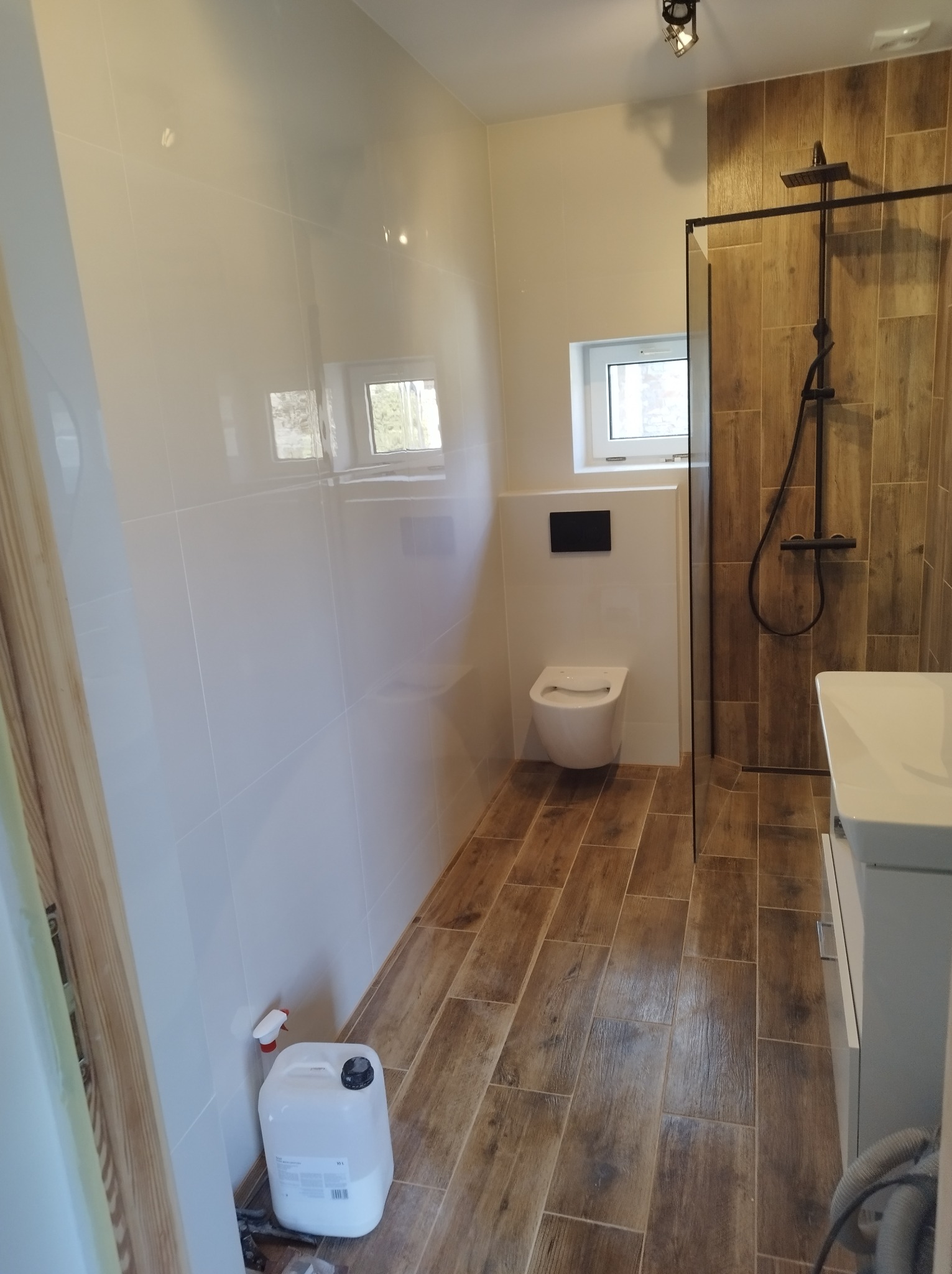 Nowoczesna łazienka z białymi płytkami ściennymi, drewnopodobną podłogą, kabiną prysznicową typu walk-in z czarną armaturą i podtynkową toaletą; widoczny pojemnik z płynem do czyszczenia...