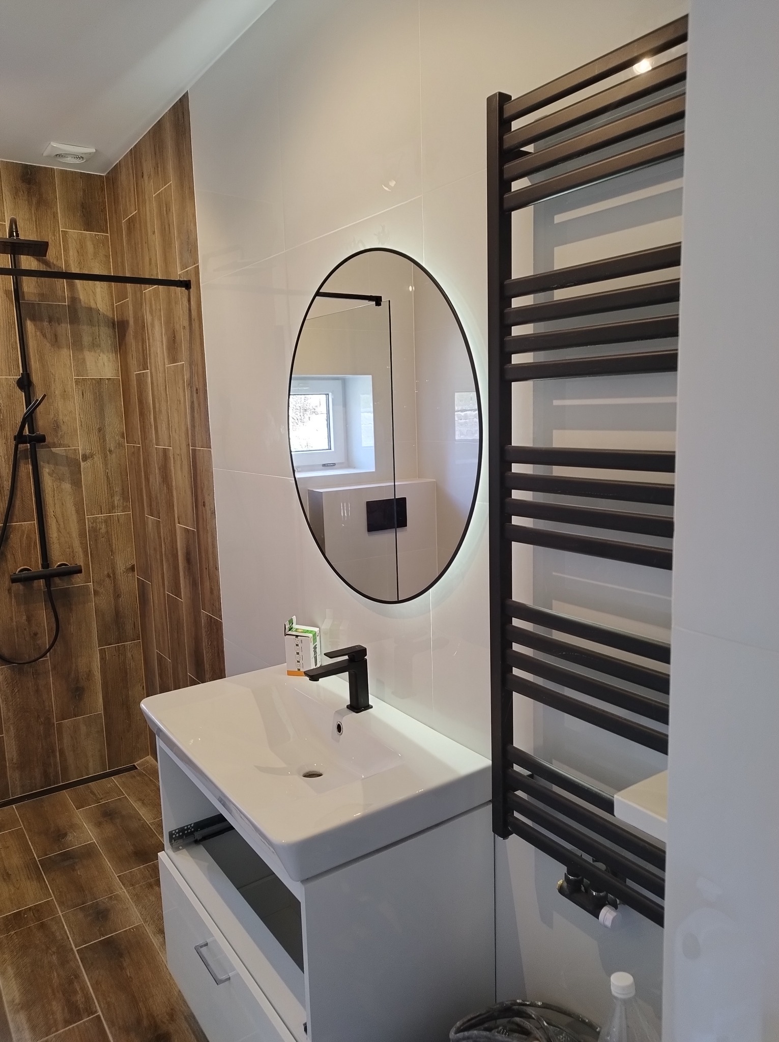 Nowoczesna łazienka z drewnopodobnymi płytkami, białymi ścianami, okrągłym lustrem z podświetleniem LED, czarną armaturą i grzejnikiem drabinkowym.