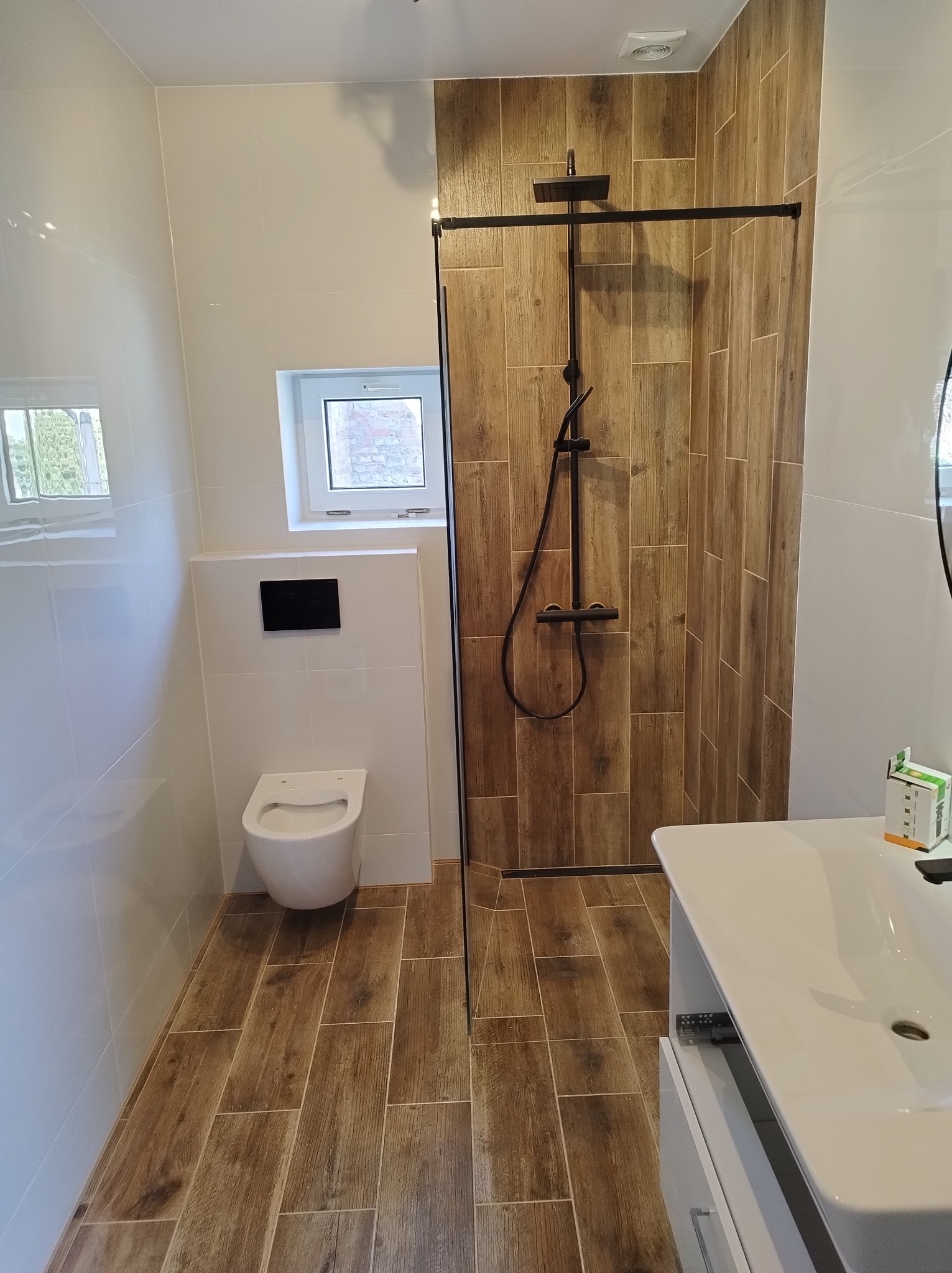 Nowoczesna łazienka z drewnopodobnymi płytkami na ścianie prysznica i podłodze, białe płytki na pozostałych ścianach, wisząca toaleta i umywalka z szafką, czarna armatura prysznicowa.