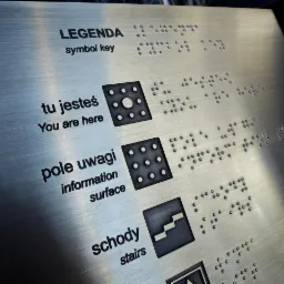 Metalowa tablica informacyjna z opisami w języku polskim i angielskim oraz alfabetem Braille'a, zawierająca symbole i napisy takie jak 'Legenda', 'Tu jesteś', 'Pole uwagi' i 'Schody'.