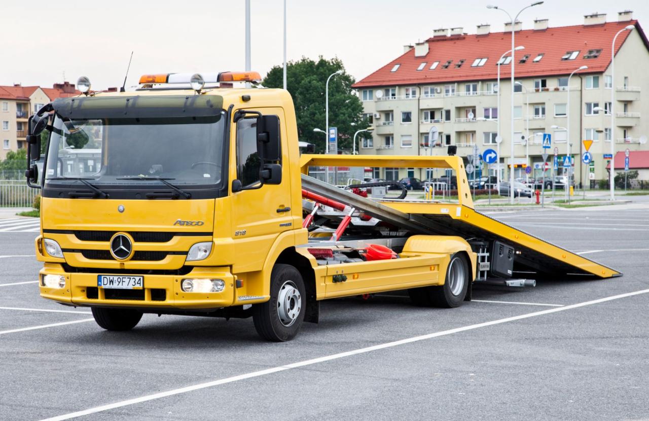 Żółta autolaweta Mercedes-Benz Atego na parkingu miejskim, z widoczną platformą załadunkową i elementami zabezpieczającymi ładunek, budynki mieszkalne w tle.