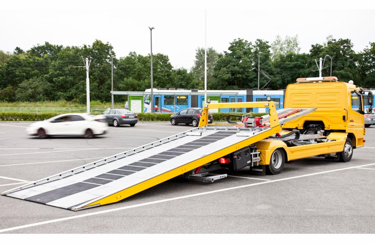 Żółta autolaweta z opuszczoną platformą na parkingu, w tle przejeżdżający tramwaj i samochody.