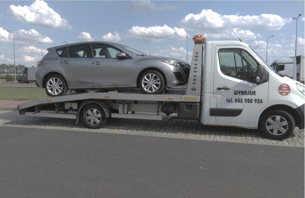 Srebrny samochód osobowy marki Mazda na platformie białej autolawety na tle parkingu i pochmurnego nieba. Na kabinie lawety widoczny napis 'WYNAJEM tel. 605 980 926' oraz informacja o cenie za dobę.