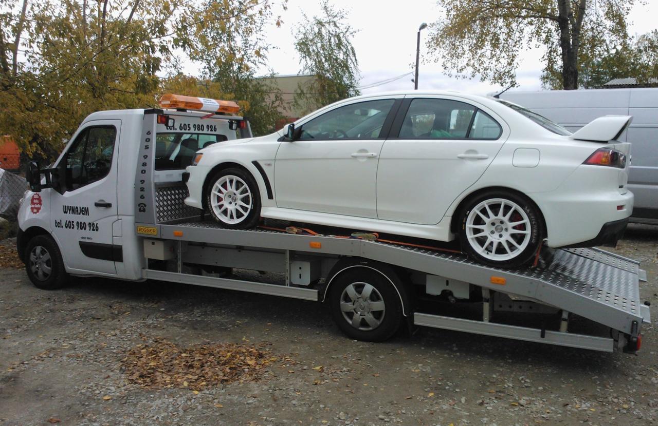 Biała laweta z białym Mitsubishi Lancer Evolution na platformie, widoczne numery telefonu do wypożyczalni lawet na karoserii, jesienne liście na ziemi.