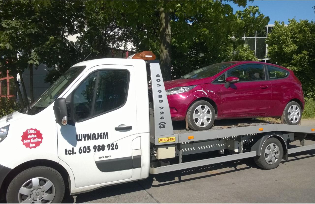 Biała laweta z uszkodzonym, czerwonym Fordem Fiesta na platformie, widoczny numer telefonu do wynajmu, tło z zielenią drzew.