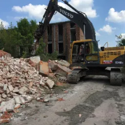 Żółta koparka Volvo EC240BLC z hydraulicznymi nożycami kruszy ceglany budynek w trakcie rozbiórki, na pierwszym planie sterta gruzu.