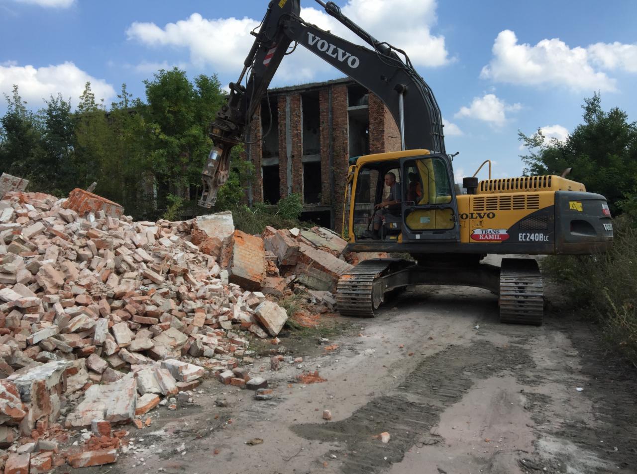 Żółta koparka Volvo EC240BLC z hydraulicznymi nożycami kruszy ceglany budynek w trakcie rozbiórki, na pierwszym planie sterta gruzu.