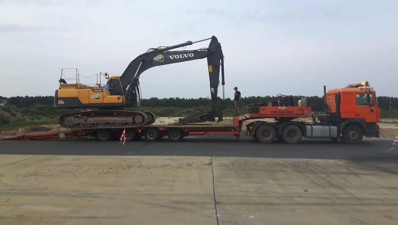 Żółto-czarna koparka Volvo EC250D transportowana na niskopodwoziowej platformie przez pomarańczową ciężarówkę MAN, widok z boku na betonowej powierzchni.